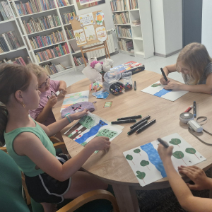Przy stole siedzą dzieci, rysują coś na kartkach,przed nimi leżą kolorowe mazaki.
