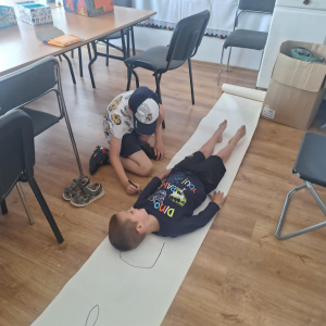 Chłopiec odrysowuje postać kolegi na dużym arkuszu papieru.