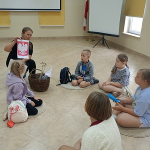 Dzieci siedzą w kręgu na podłodze, pani bibliotekarka trzyma w ręku godło Polski.