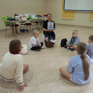 Dzieci siedzą w kręgu na podłodze, pani bibliotekarka trzyma w ręku książkę.