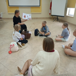 Dzieci siedzą w kręgu na podłodze, pani bibliotekarka pokazuje ilustracje w książce.