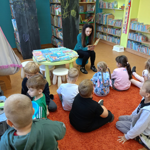 Na małym krzesełku siedzi kobieta,czyta książkę przed nią na dywanie siedzą dzieci.