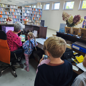 Przy biurku siedzi bibliotekarka za nią stoją dzieci podają jej książki.