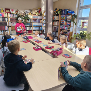 Przy stole siedzą dzieci, przed nimi stoi prowadząca, trzyma w ręku książkę, na stole leżą różne przybory.