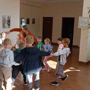 Dzieci wraz z prowadzącą stoją ustawieni w kole, trzymają się za ręce.