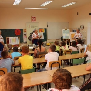 Dzieci siedzące w ławkach. Trzy kobiety w strojach ludowych