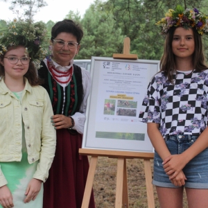 Prowadząca i dwie dziewczynki po bokach w wiankach na głowie. Obok prowadzącej plakat.