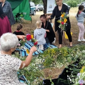 Uczestnicy siedzą na rozłożonych kocach i wiją wianki.