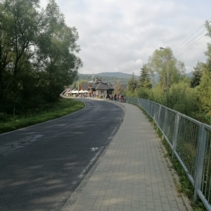 Droga obok znajdującego się w pobliżu lasu oraz z  zabudowaniami w tle 