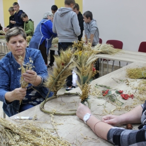 Kobieta o blond włosach zawija sznurkiemm zioła na stelaż. Inna kobieta trzyma kłosy zboża w ręku.