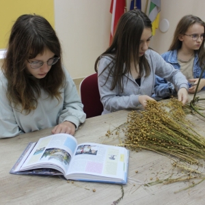 Trzy dziewczynki siedza przy stole. Na stole leżą książka, zioła i wienieć dożylkowy z gałęzi i ziół.