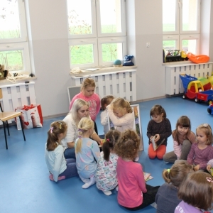 Dzieci siedzą w kręgu na podłodze. Dziewczynka trzyma portret z podobizną Konopnickiej.